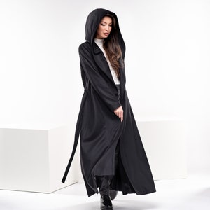 May include: A long black hooded coat with a tie belt. The coat is worn by a person with long brown hair. The person is standing on a white surface.