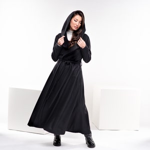May include: A black hooded long coat with a wrap style and a belt. The coat is worn by a person with long brown hair. The person is standing on a white background.