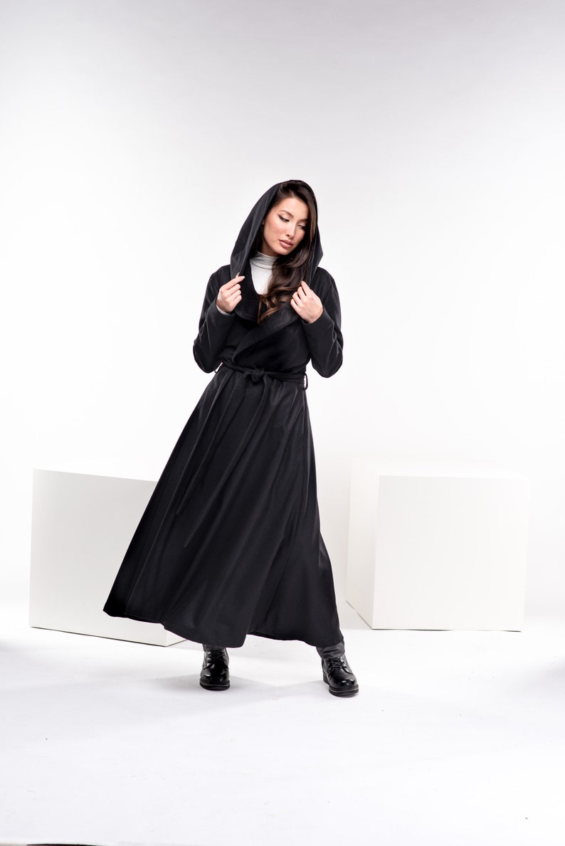 May include: A black hooded long coat with a wrap style and a belt. The coat is worn by a person with long brown hair. The person is standing on a white background.