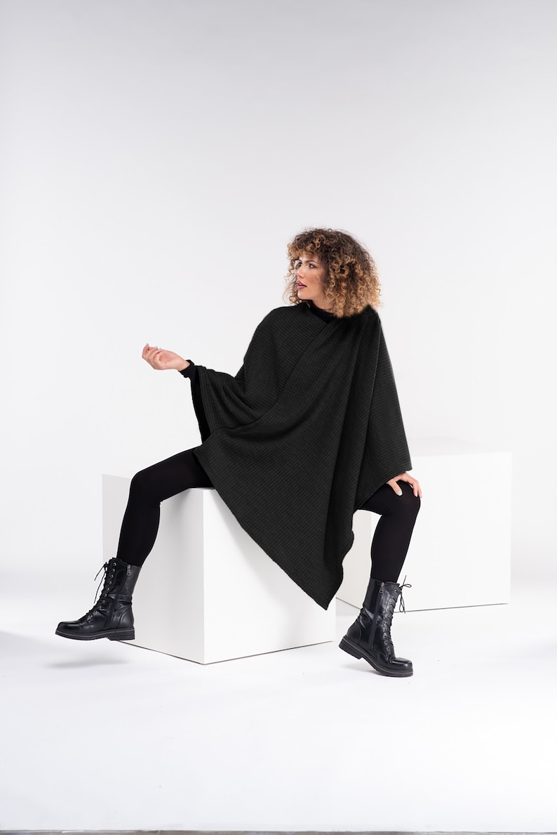 May include: A woman wearing a black knit poncho and black leggings sits on a white cube. She is wearing black lace-up boots.