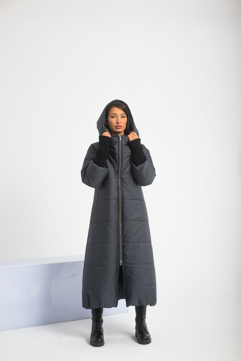 May include: A long, dark gray puffer coat with a zipper closure and a hood. The coat has a quilted design and is worn by a person with black boots.