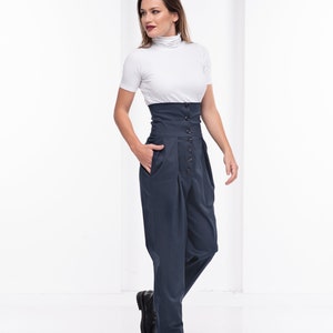 Pu&ograve; includere: Una donna indossa un top bianco a maniche corte con collo alto e pantaloni blu navy a vita alta. I pantaloni hanno una chiusura con bottoni e tasche. Scarpe in pelle nera completano l'outfit. Lo sfondo &egrave; bianco.