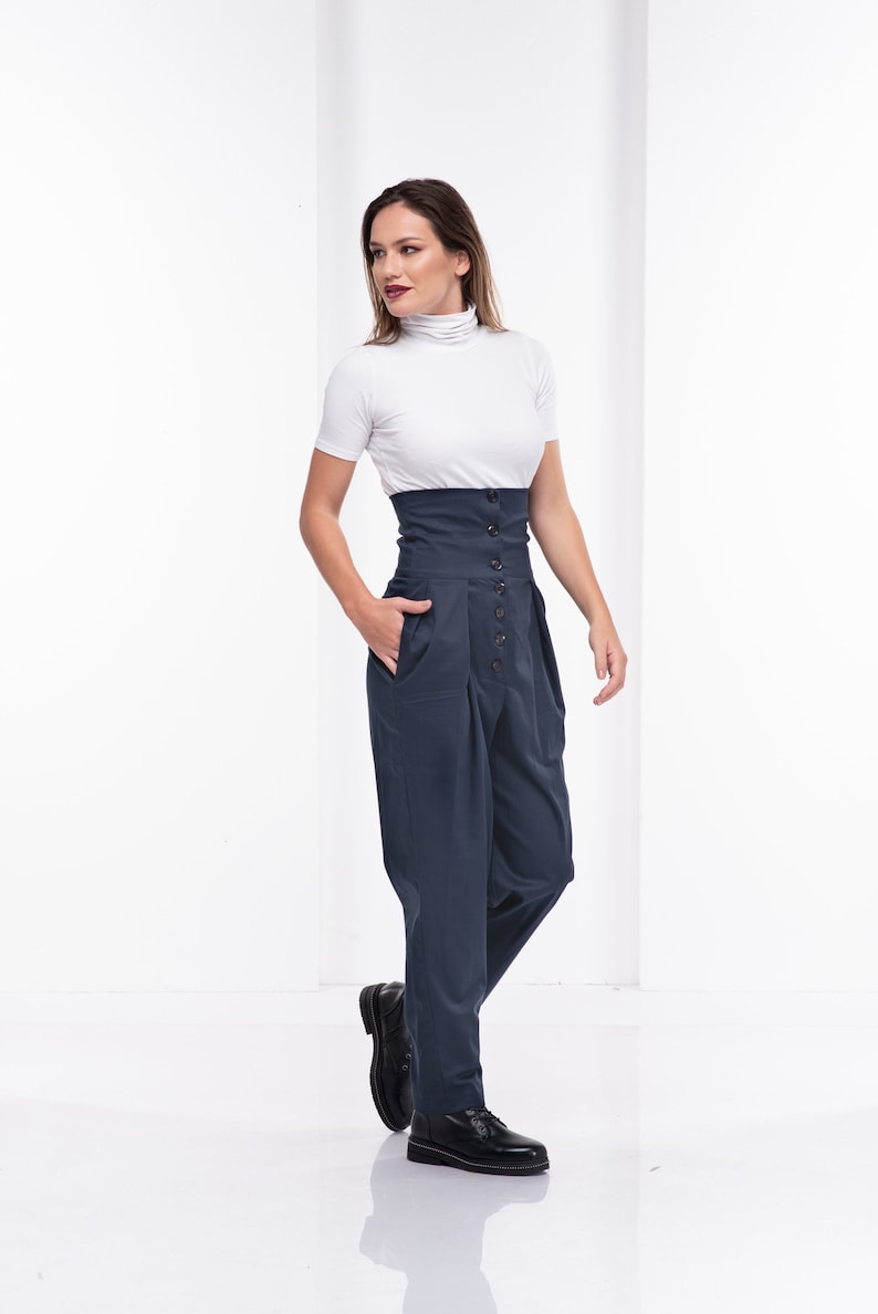 Pu&ograve; includere: Una donna indossa un top bianco a maniche corte con collo alto e pantaloni blu navy a vita alta. I pantaloni hanno una chiusura con bottoni e tasche. Scarpe in pelle nera completano l'outfit. Lo sfondo &egrave; bianco.