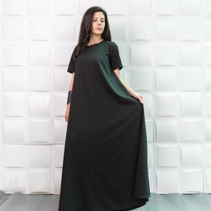 K&ouml;nnte beinhalten: Eine Frau tr&auml;gt ein langes, schwarzes Maxikleid mit kurzen &Auml;rmeln. Das Kleid hat einen schlichten, lockeren Schnitt und reicht ihr bis zu den Kn&ouml;cheln. Sie steht vor einer wei&szlig;en Wand mit einem strukturierten Muster.