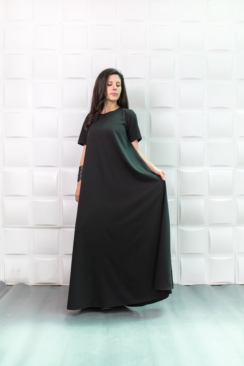 K&ouml;nnte beinhalten: Eine Frau tr&auml;gt ein langes, schwarzes Maxikleid mit kurzen &Auml;rmeln. Das Kleid hat einen schlichten, lockeren Schnitt und reicht ihr bis zu den Kn&ouml;cheln. Sie steht vor einer wei&szlig;en Wand mit einem strukturierten Muster.