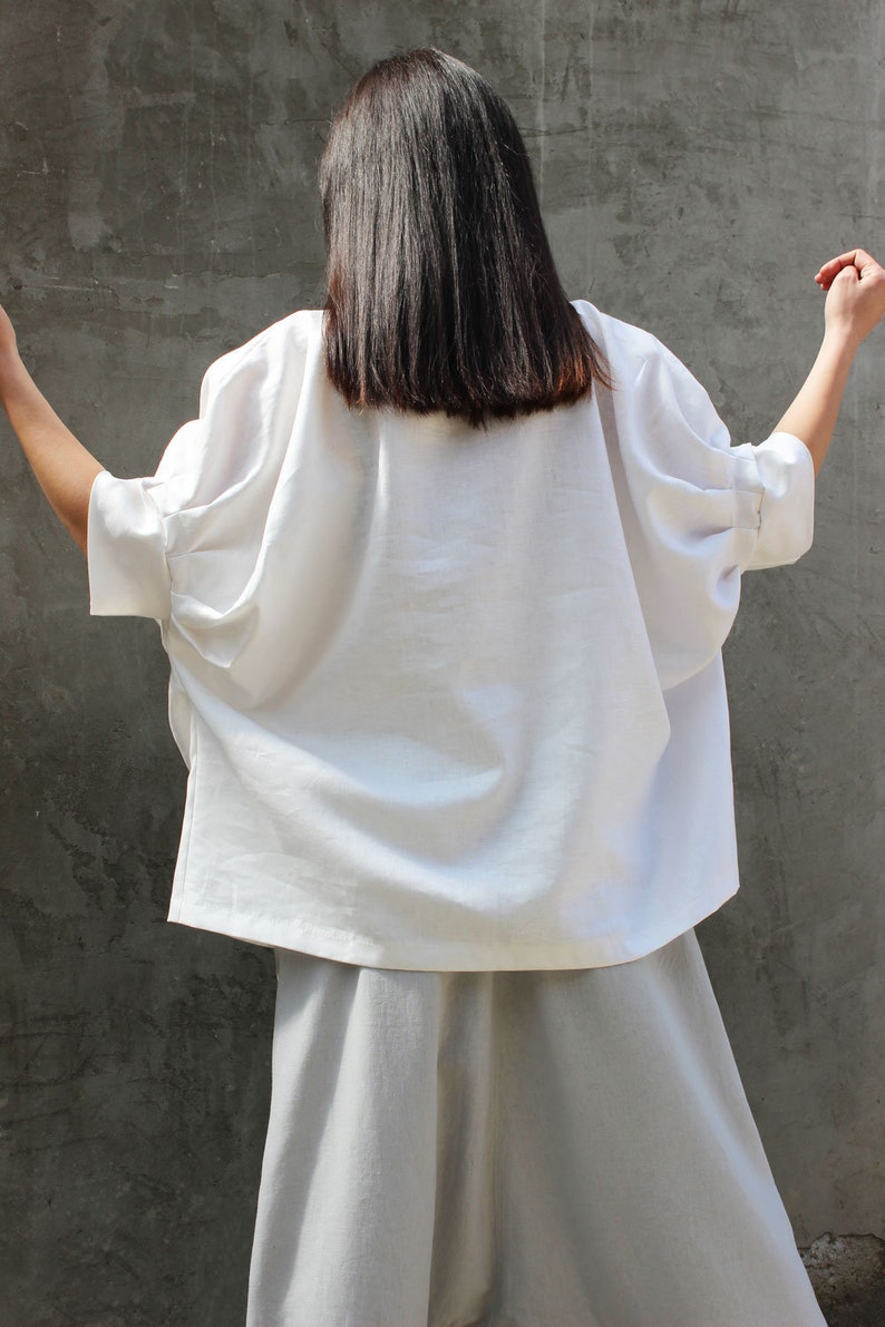 May include: A white linen blouse with a relaxed fit and long sleeves. The blouse is worn with a pair of light gray linen pants.