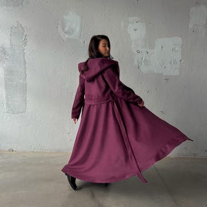 May include: A long, flowing, hooded coat in a deep burgundy color. The coat features a belted waist and a full skirt, creating a dramatic silhouette. The fabric appears to be a soft, draping material.