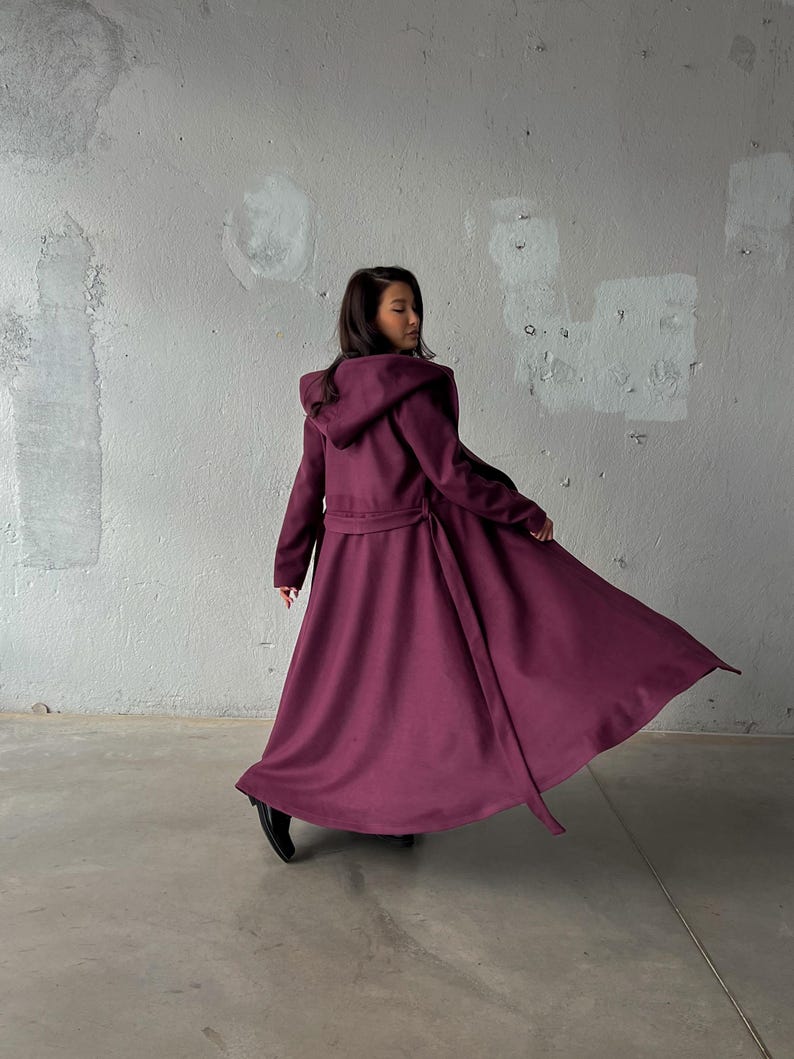 May include: A long, flowing, hooded coat in a deep burgundy color. The coat features a belted waist and a full skirt, creating a dramatic silhouette. The fabric appears to be a soft, draping material.
