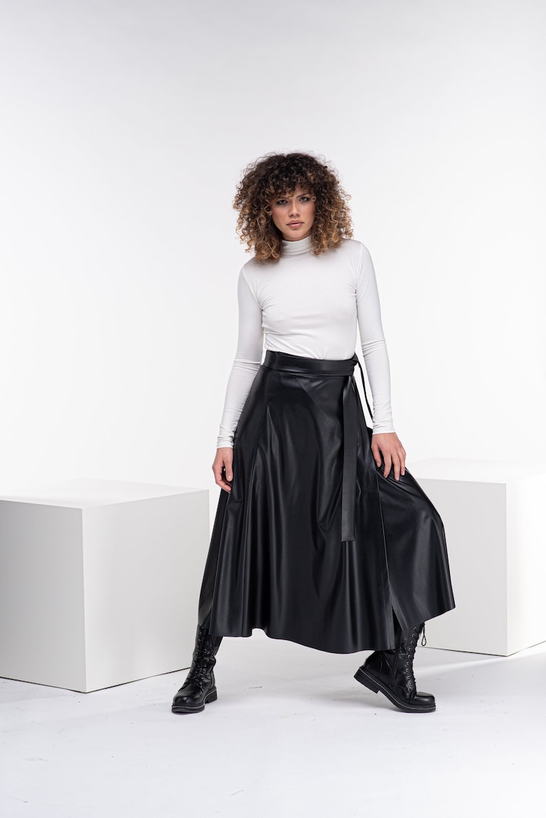 May include: A woman wearing a white turtleneck top and a black faux leather wrap skirt. She is standing on a white platform and wearing black boots.