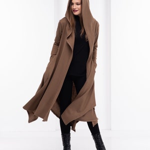 May include: A long brown hooded cardigan with a draped front and long sleeves. The cardigan is worn over a black turtleneck and black pants. The person is wearing black boots.