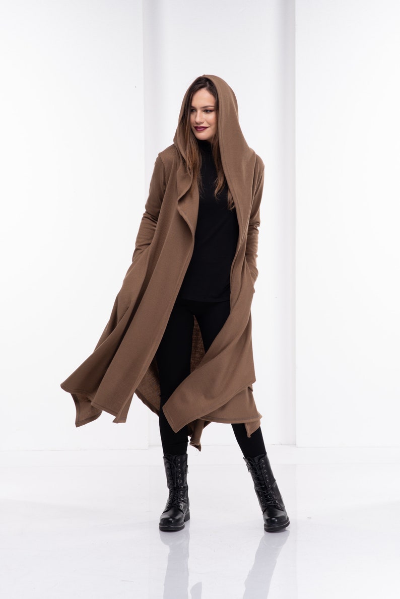 May include: A long brown hooded cardigan with a draped front and long sleeves. The cardigan is worn over a black turtleneck and black pants. The person is wearing black boots.