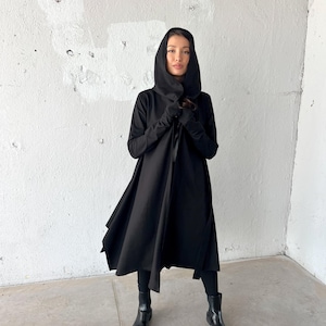 May include: A black hooded coat with long sleeves and a flowing skirt. The coat is open in the front and worn with black leggings and black ankle boots. The background is a white wall.