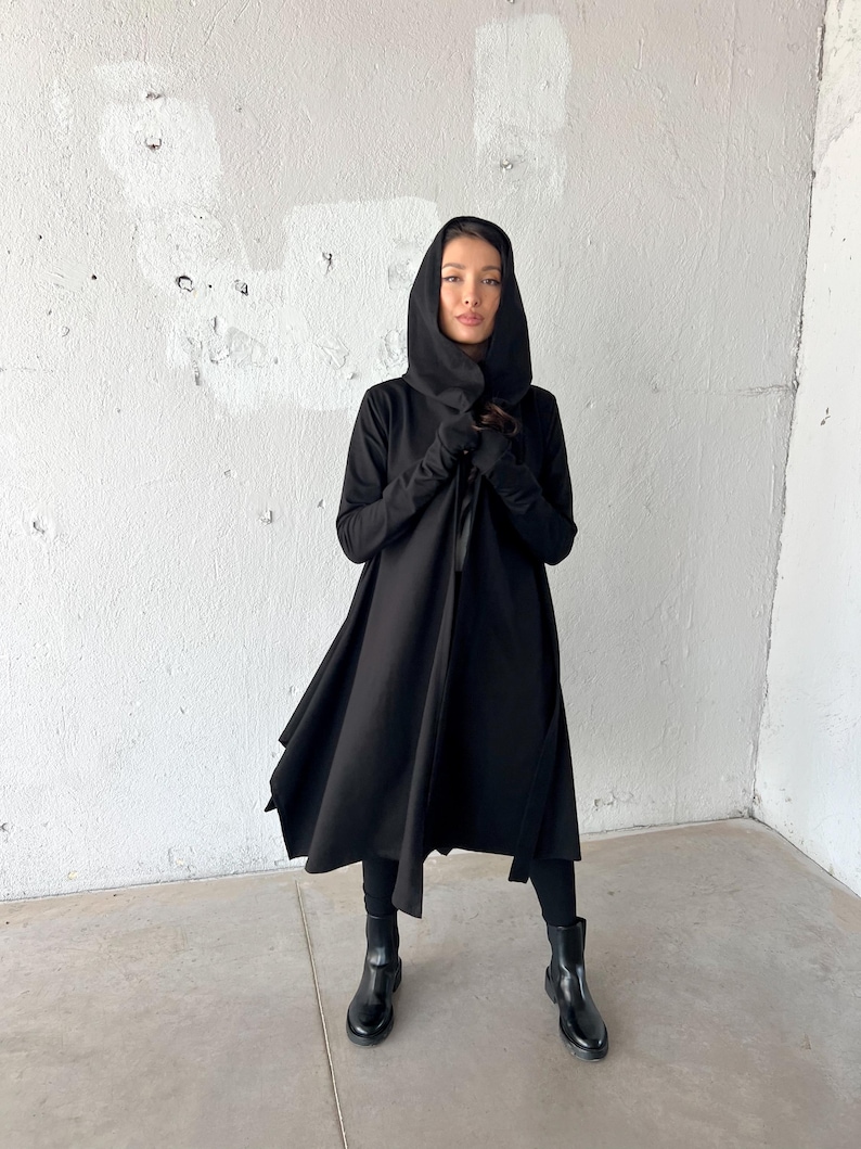 May include: A black hooded coat with long sleeves and a flowing skirt. The coat is open in the front and worn with black leggings and black ankle boots. The background is a white wall.