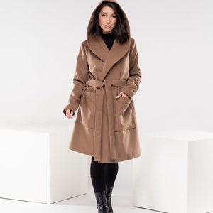 May include: A brown hooded wrap coat with a belt and pockets. The coat is worn by a person with black leggings and black boots.
