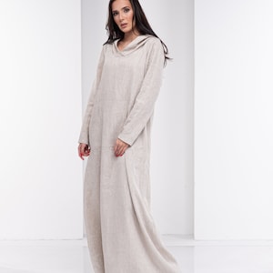 May include: A long, beige linen dress with a hood. The dress has a relaxed fit and a flowing silhouette. The model is wearing the dress with a natural, effortless look.