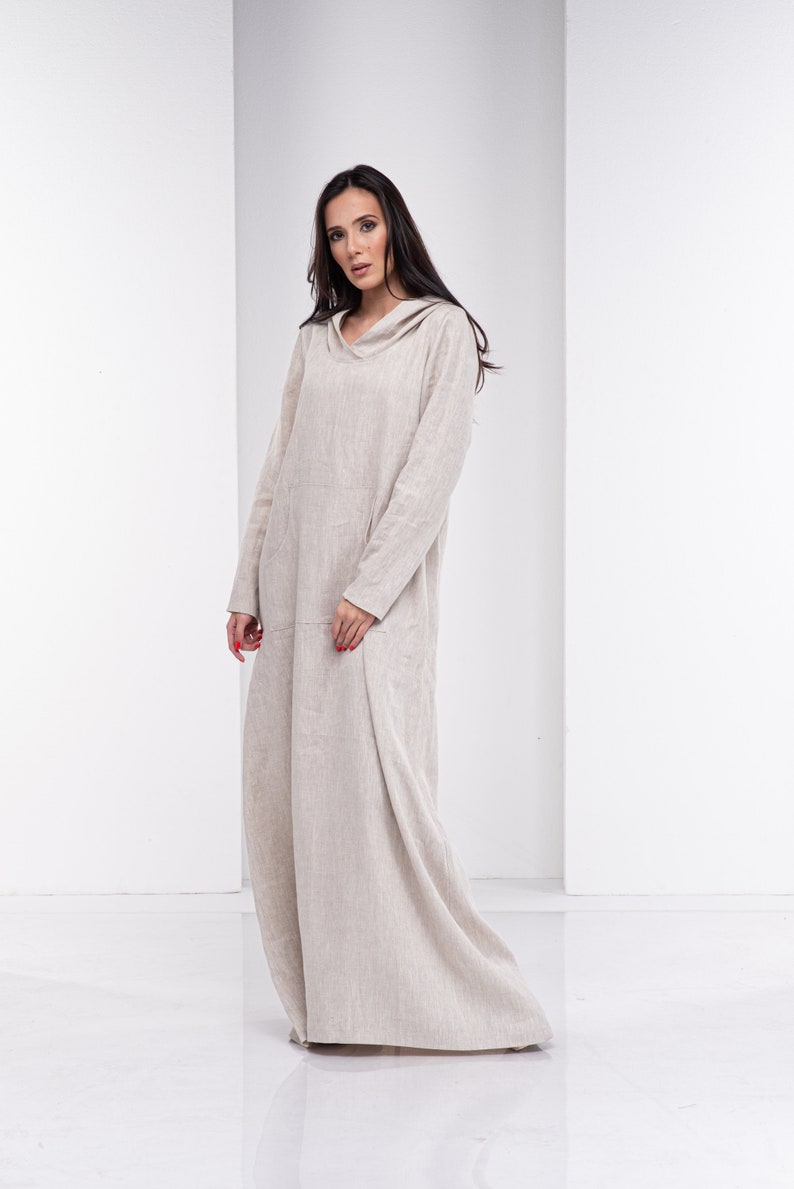 May include: A long, beige linen dress with a hood. The dress has a relaxed fit and a flowing silhouette. The model is wearing the dress with a natural, effortless look.