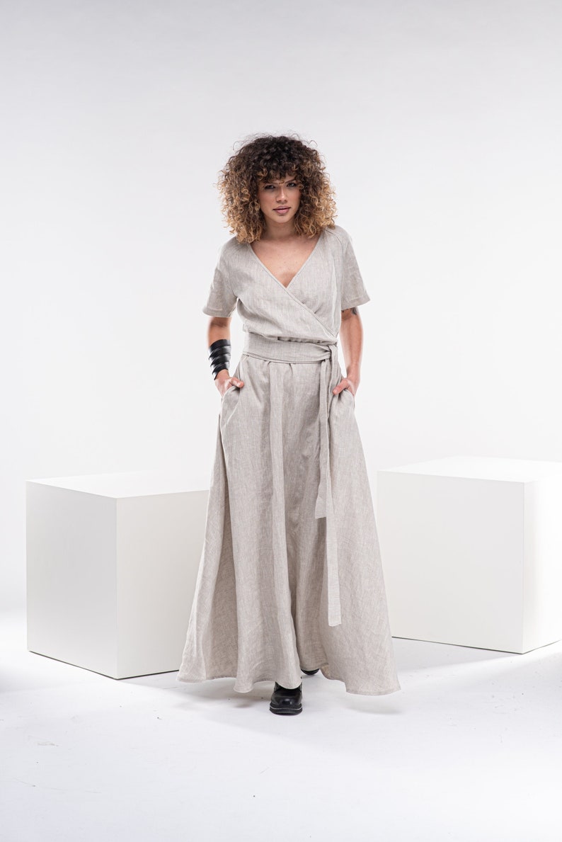 May include: A woman wearing a light beige linen wrap dress with a long skirt and short sleeves. She is standing on a white platform with her hands in her pockets.