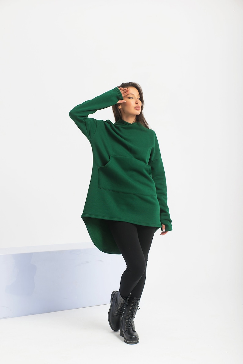 May include: A woman wearing a dark green hooded sweatshirt with a long, asymmetrical hemline and black leggings. She is wearing black lace-up boots.