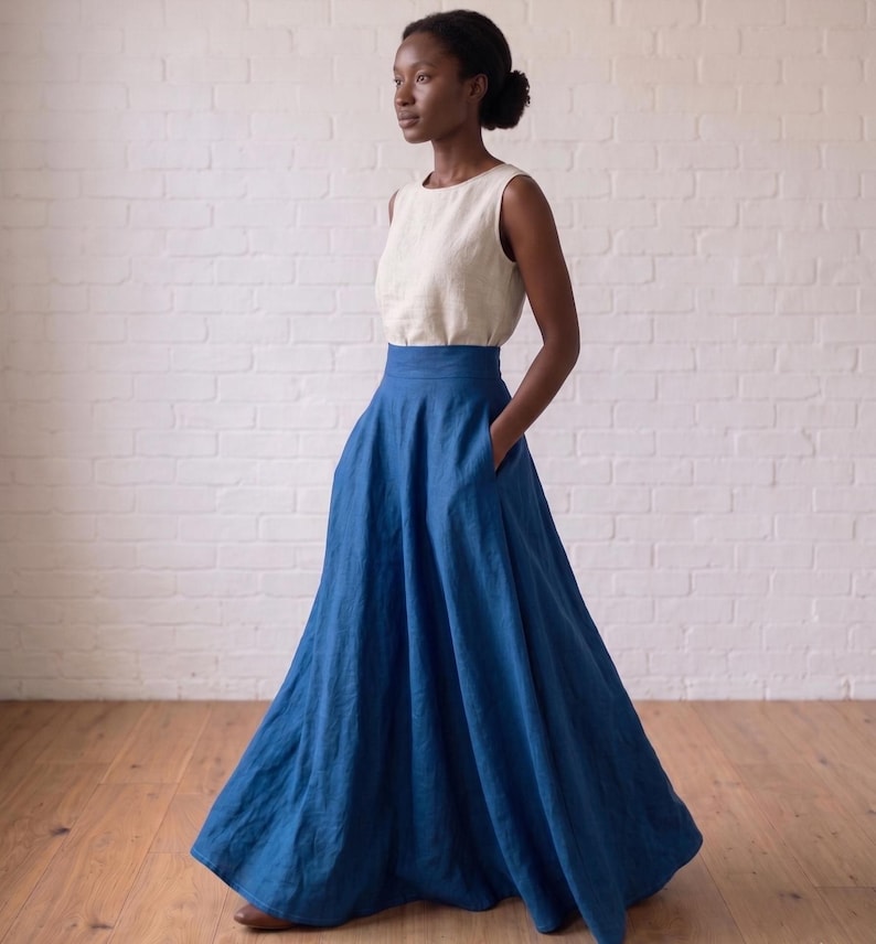 May include: A sleeveless cream-colored top paired with a high-waisted, floor-length blue skirt. The skirt features a wide waistband, a full, flowing silhouette, and side pockets. The ensemble is composed of a solid-colored, textured fabric.