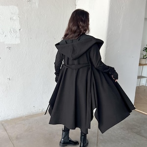 May include: A long black hooded coat with a flowing, asymmetrical hemline. The coat features a belted waist and long sleeves. The person is wearing black boots. The coat is made of a soft, flowing fabric.