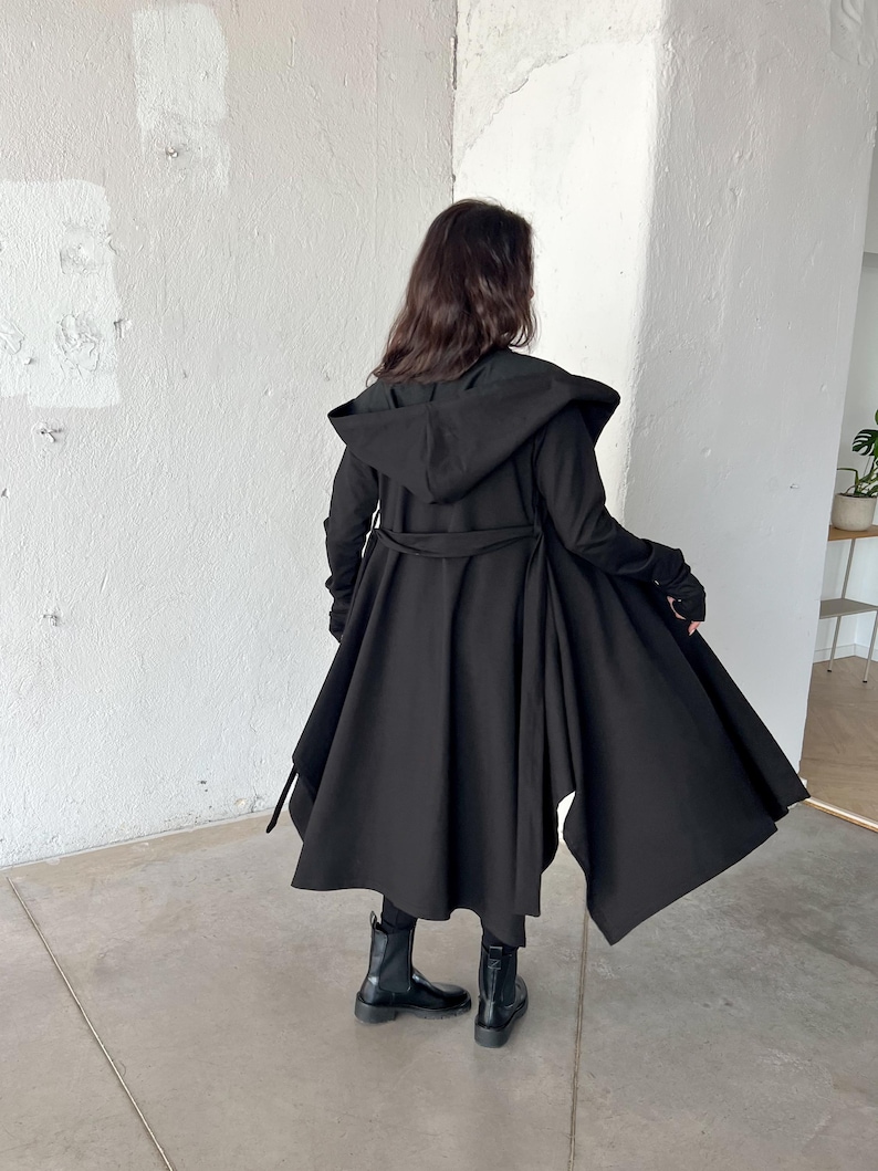 May include: A long black hooded coat with a flowing, asymmetrical hemline. The coat features a belted waist and long sleeves. The person is wearing black boots. The coat is made of a soft, flowing fabric.