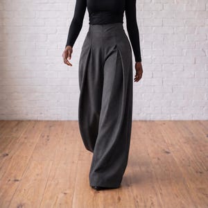 Op de afbeelding: Hoge taille broek in antracietgrijs met wijde pijpen, diepe plooien aan de voorzijde en een gestructureerde tailleband. De broek wordt gecombineerd met een zwart shirt met lange mouwen voor een strakke, getailleerde uitstraling met een lange val.