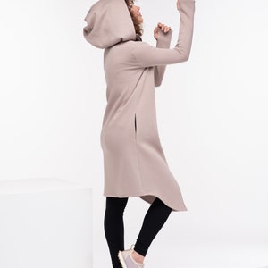 May include: A woman wearing a long, beige hooded sweatshirt with a high neckline and long sleeves. She is wearing black leggings and light pink sneakers. The sweatshirt has a long, asymmetrical hemline and side slits.