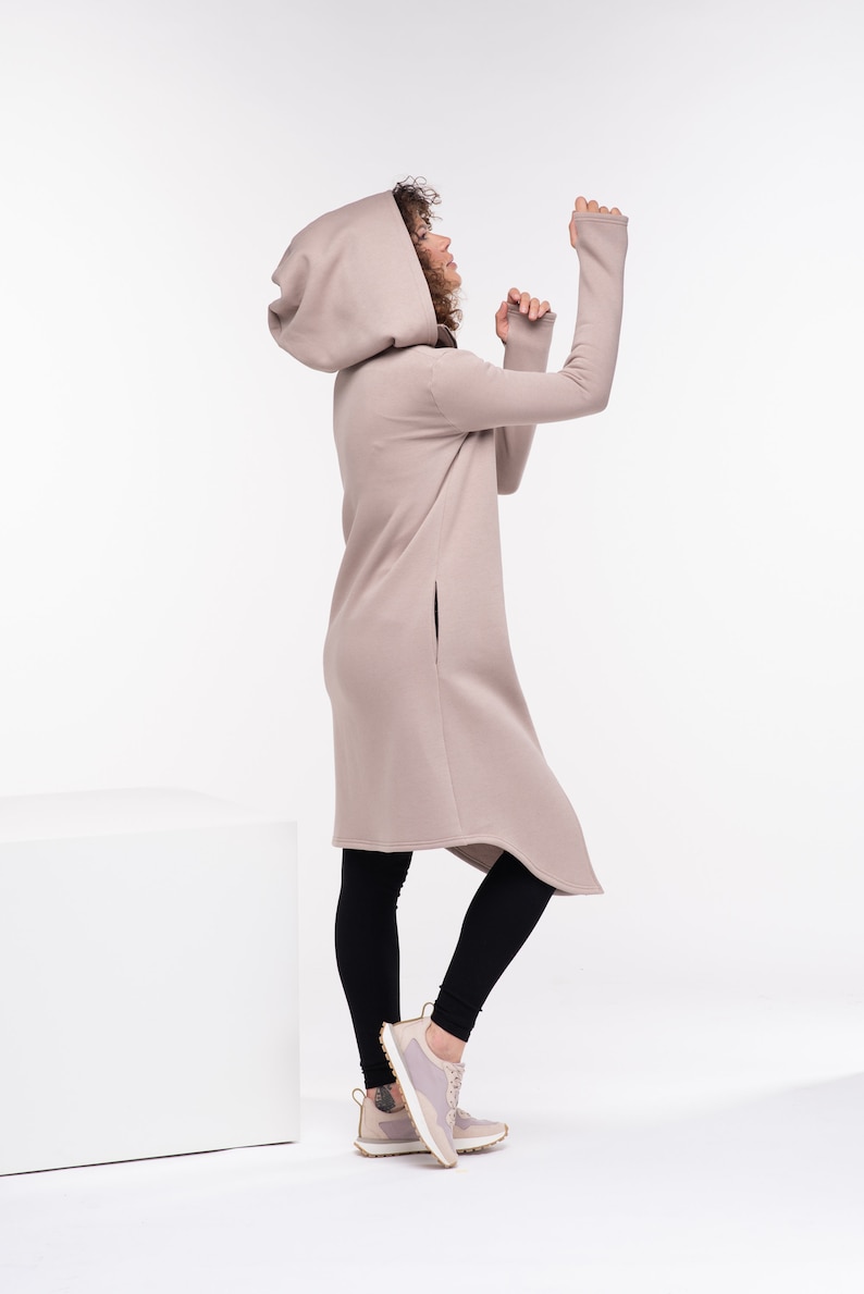 May include: A woman wearing a long, beige hooded sweatshirt with a high neckline and long sleeves. She is wearing black leggings and light pink sneakers. The sweatshirt has a long, asymmetrical hemline and side slits.