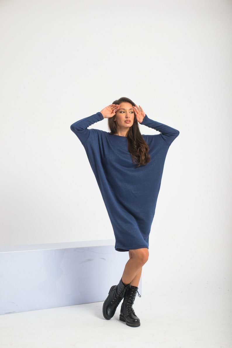 May include: A woman wearing a long, loose-fitting, navy blue dress with long sleeves. She is wearing black lace-up boots and has her hands on her head.