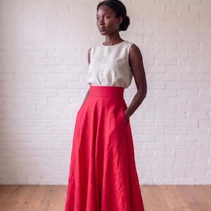 May include: A sleeveless beige top with a round neckline paired with a high-waisted, floor-length red skirt. The skirt features a wide waistband and side pockets, creating a full, flowing silhouette that reaches the floor in a solid red color.