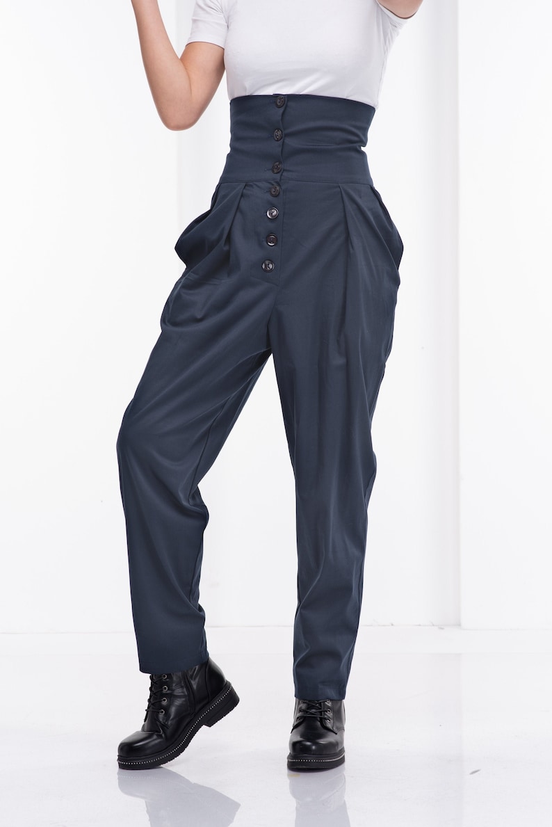 Pu&ograve; includere: Pantaloni blu navy a vita alta con chiusura a bottoni e parte anteriore a pieghe. I pantaloni hanno un'ampia fascia in vita e sono abbinati a stivali neri. La modella indossa anche una maglietta bianca.