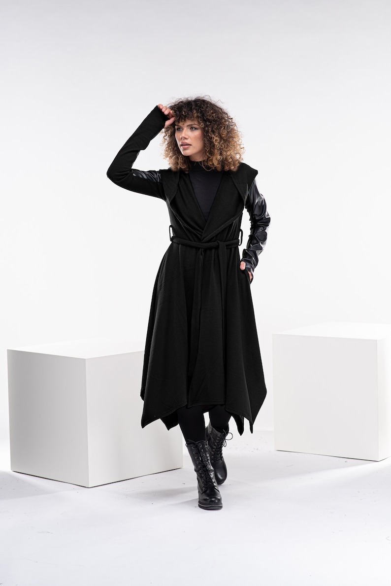 May include: A black hooded coat with leather sleeves and a tie belt. The coat has a long, flowing silhouette and is worn by a person with curly hair.
