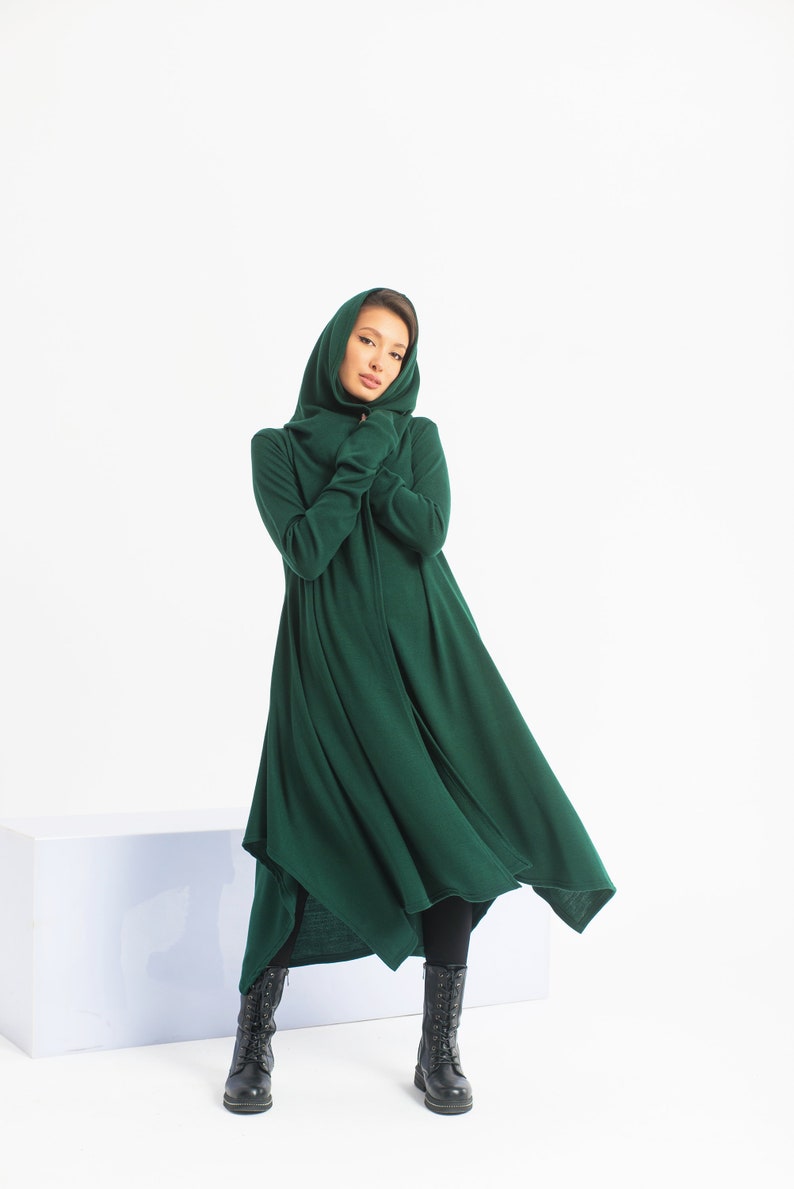 May include: A long, dark green hooded dress with a flowing skirt. The dress is made of a soft, comfortable fabric and has a relaxed fit. The model is wearing black boots.