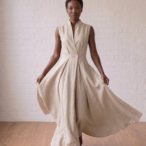 May include: A sleeveless beige dress with a high collar, a deep V-neckline, and a single button closure at the waist. The garment features a fitted bodice that transitions into a full, pleated skirt that flares outward at the hemline.