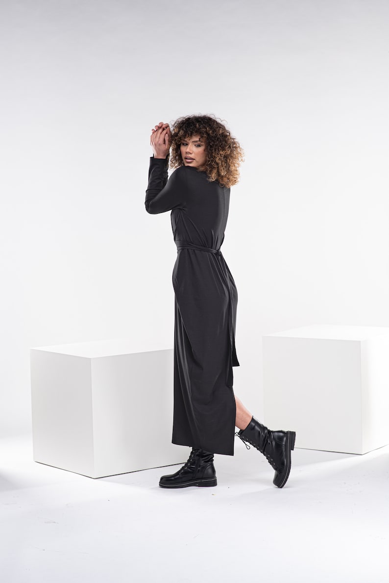 May include: A woman wearing a black maxi dress with a side slit and a belt. She is wearing black boots and has curly brown hair.