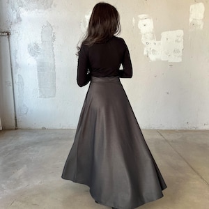 May include: A black long-sleeved shirt with a black skirt. The skirt is a-line and falls to the ankles. The person is standing in front of a white wall.