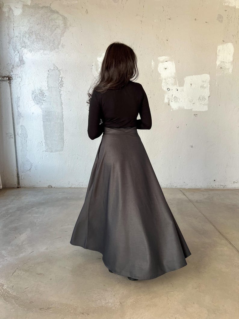 May include: A black long-sleeved shirt with a black skirt. The skirt is a-line and falls to the ankles. The person is standing in front of a white wall.