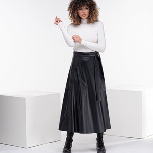 May include: A woman wearing a white turtleneck sweater and a black faux leather wrap skirt. She is wearing black boots and has curly brown hair.