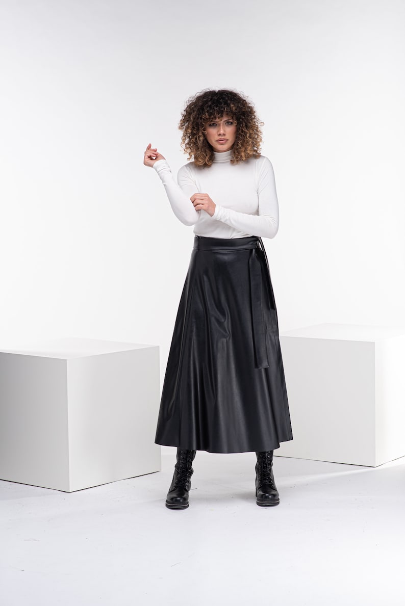 May include: A woman wearing a white turtleneck sweater and a black faux leather wrap skirt. She is wearing black boots and has curly brown hair.