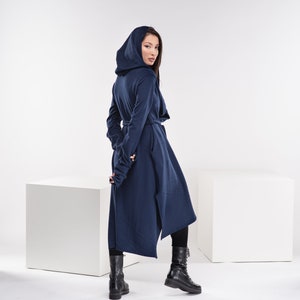 May include: A long, navy blue hooded cardigan with a long, asymmetrical hemline. The cardigan has a tie belt at the waist and long sleeves with thumbholes. The model is wearing black boots.