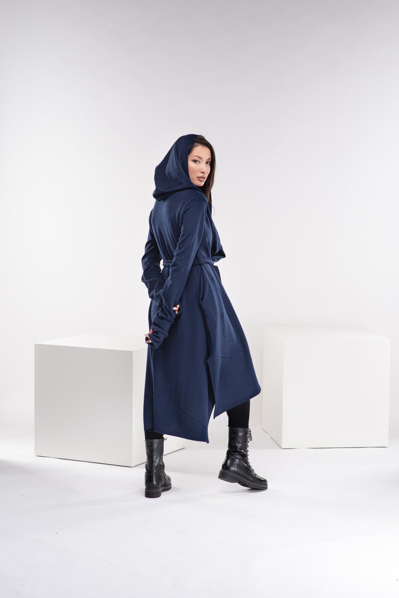 May include: A long, navy blue hooded cardigan with a long, asymmetrical hemline. The cardigan has a tie belt at the waist and long sleeves with thumbholes. The model is wearing black boots.