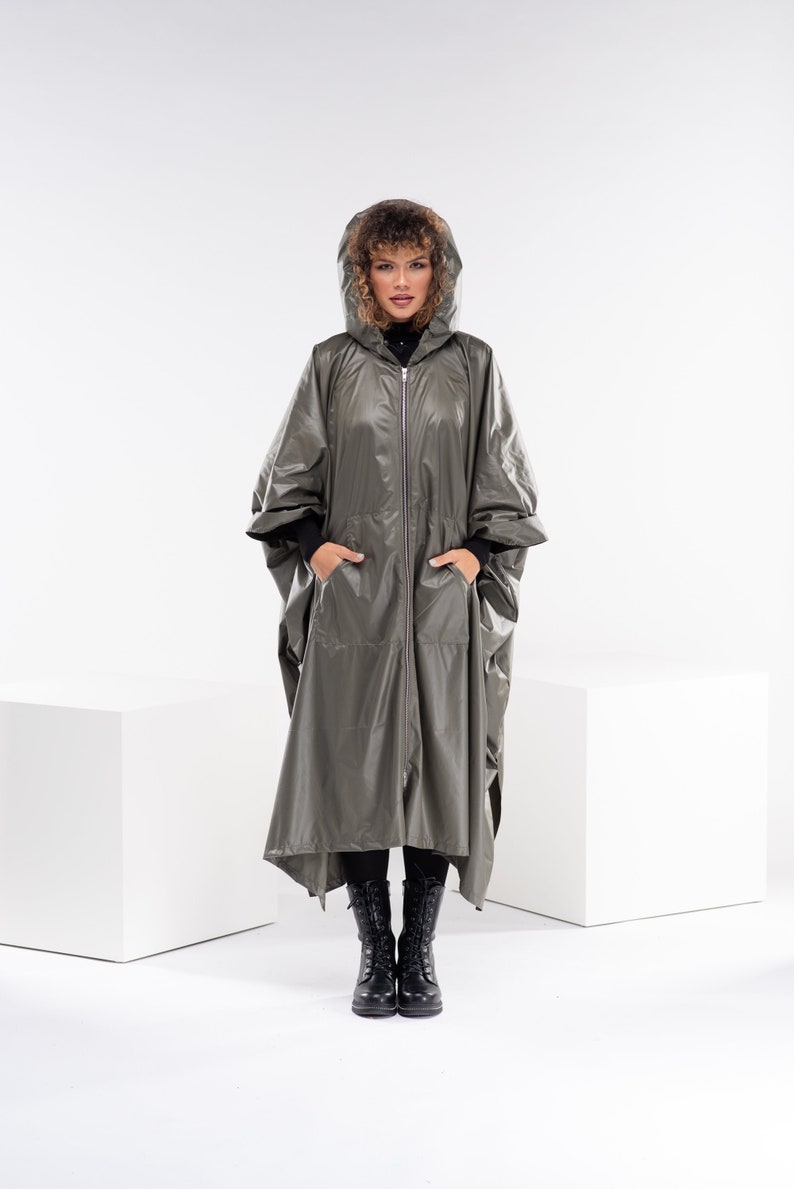 May include: A woman wearing a long, hooded, olive green raincoat with a zipper closure. She is standing on a white surface and wearing black boots.