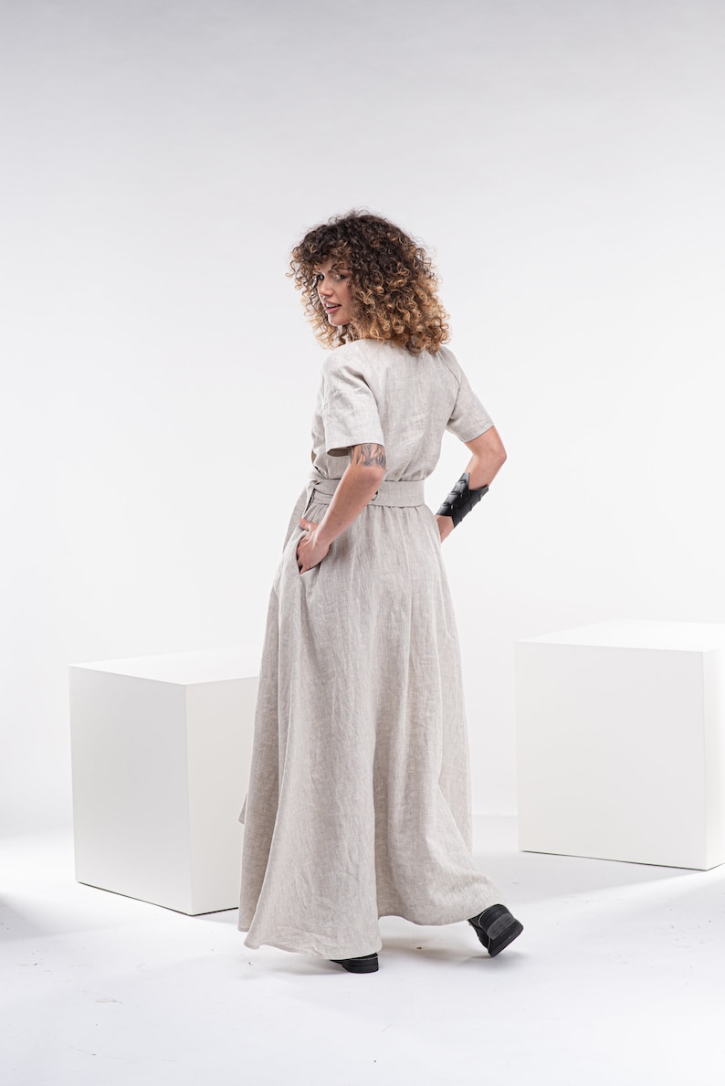 May include: A woman wearing a light beige linen dress with a black belt and black wrist cuffs. The dress has a loose fit and a flowing skirt. She is standing on a white background.