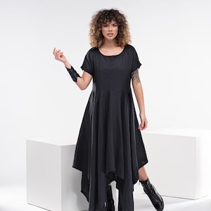 May include: A woman wearing a black asymmetrical dress with short sleeves and black boots. She is standing in front of a white background.