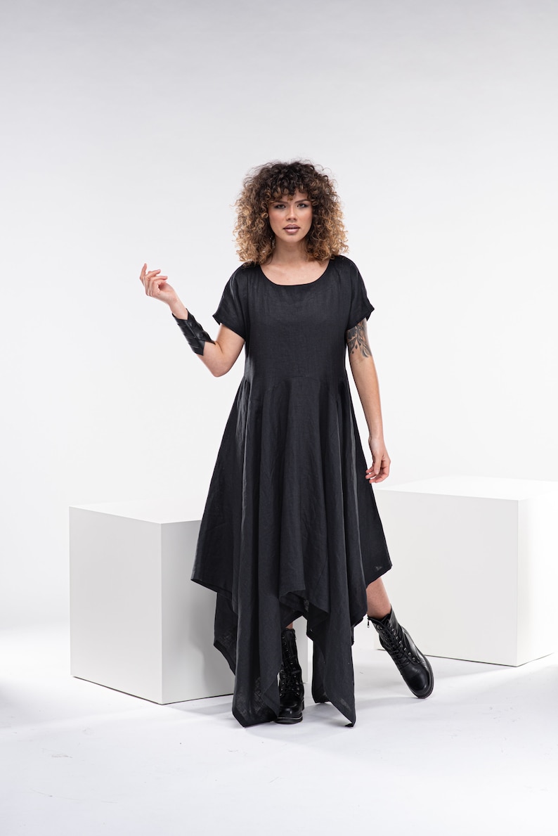 May include: A woman wearing a black asymmetrical dress with short sleeves and black boots. She is standing in front of a white background.