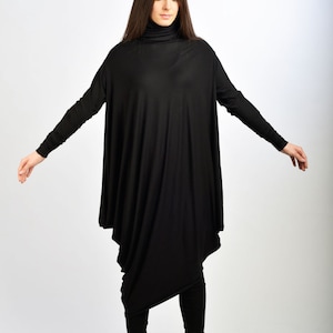 Pode incluir: Uma mulher vestindo um vestido túnica preto de manga comprida com gola alta e design drapeado. Ela está usando leggings pretos e botas de salto alto pretas.