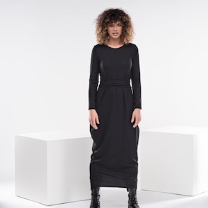 May include: A black maxi dress with long sleeves and a tie at the waist. The dress is made of a soft, flowing fabric and has a relaxed fit. The model is wearing black boots.