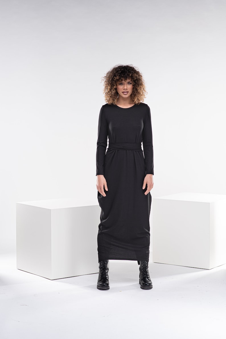 May include: A black maxi dress with long sleeves and a tie at the waist. The dress is made of a soft, flowing fabric and has a relaxed fit. The model is wearing black boots.