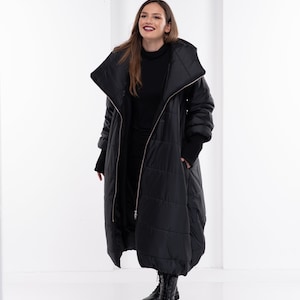 May include: A black puffer coat with a large collar and a zip closure. The coat is long and has a relaxed fit. The model is wearing black boots.