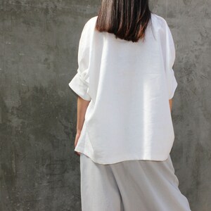 May include: A white linen shirt with a relaxed fit and three-quarter sleeves. The shirt is worn over a pair of light gray linen pants.